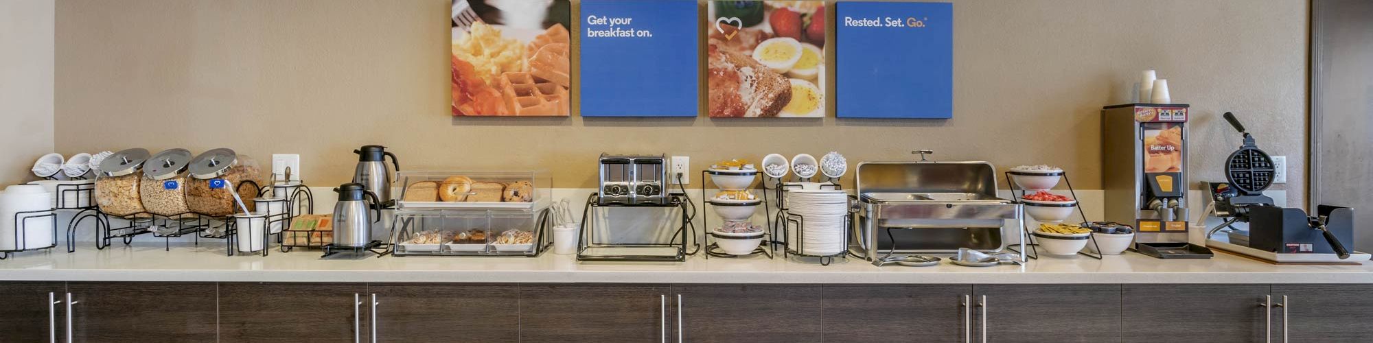 A breakfast buffet setup with various food items and appliances, including a waffle maker, coffee machine, and food dispensers, on a counter.