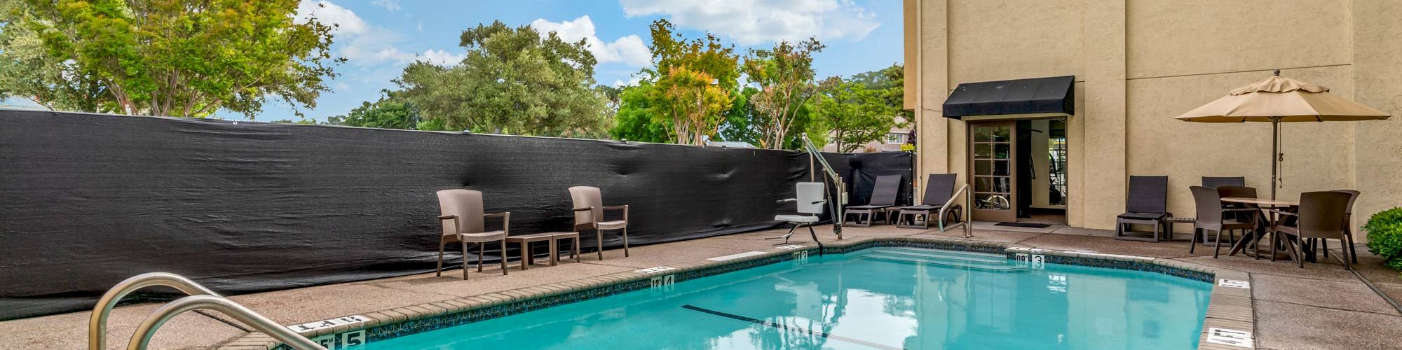 An outdoor swimming pool area with lounge chairs, umbrellas, and a building in the background under a partly cloudy sky.