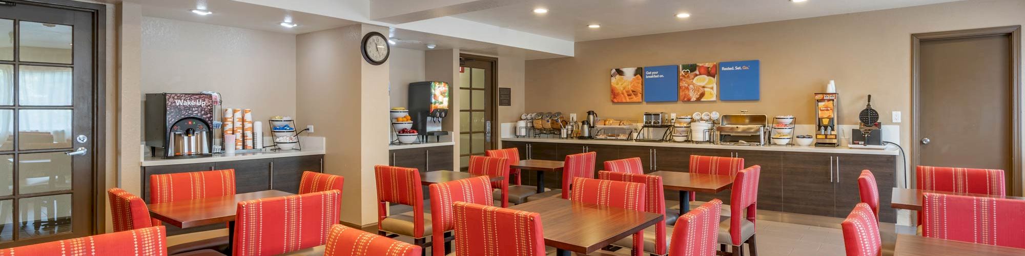 The image shows a dining area with red chairs, wooden tables, and a buffet counter with food and beverage amenities in the background.