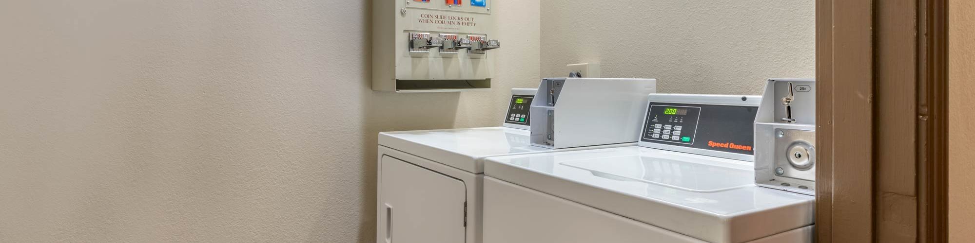 The image shows a small laundry room with a washing machine, a detergent dispensing machine, and a dryer under bright lighting.