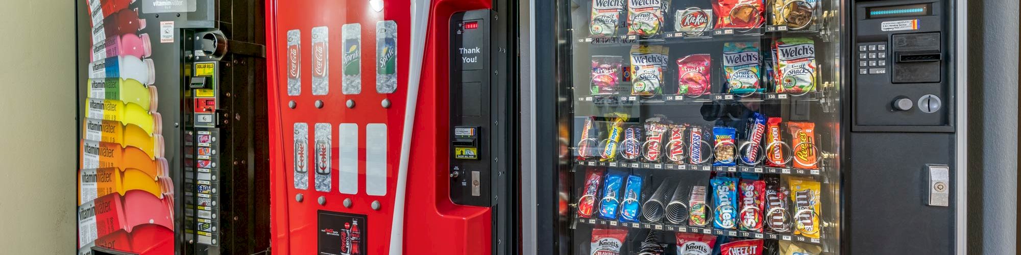 The image shows three vending machines: a soda machine with various beverages, a Coca-Cola machine, and a machine with assorted snacks and chips.