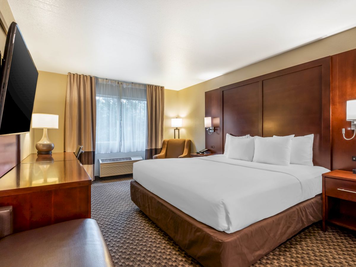 The image shows a hotel room with a large bed, TV, armchair, desk, lamps, and a window with curtains.