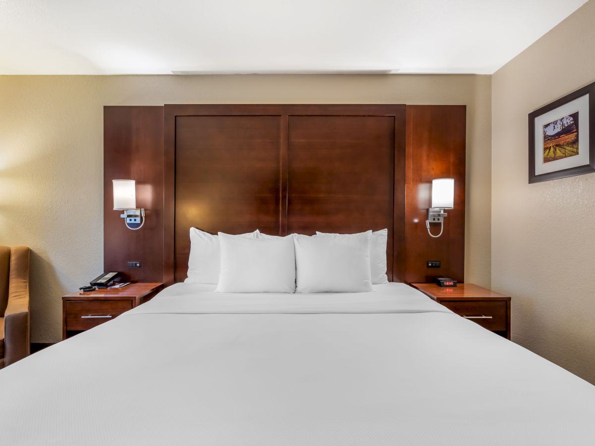 A neatly made bed with white linens, two wall-mounted lamps, wooden headboard, nightstands with phones, and framed pictures on the wall.