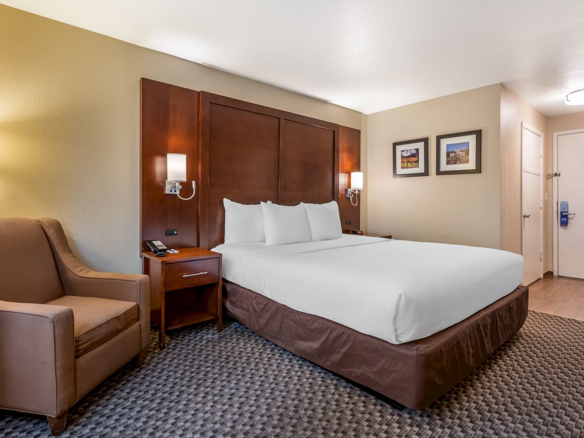 The image shows a neatly made bed in a hotel room with modern furniture, including a chair, side table, and lamps, with framed art on the wall.