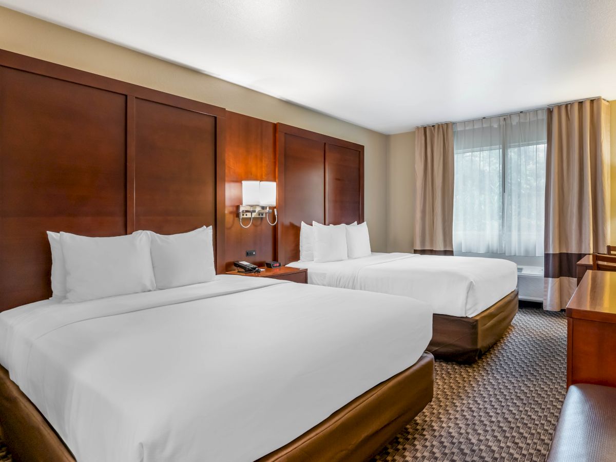 A neatly arranged hotel room with two double beds, a window with curtains, a wall lamp, and a wooden desk and chair.