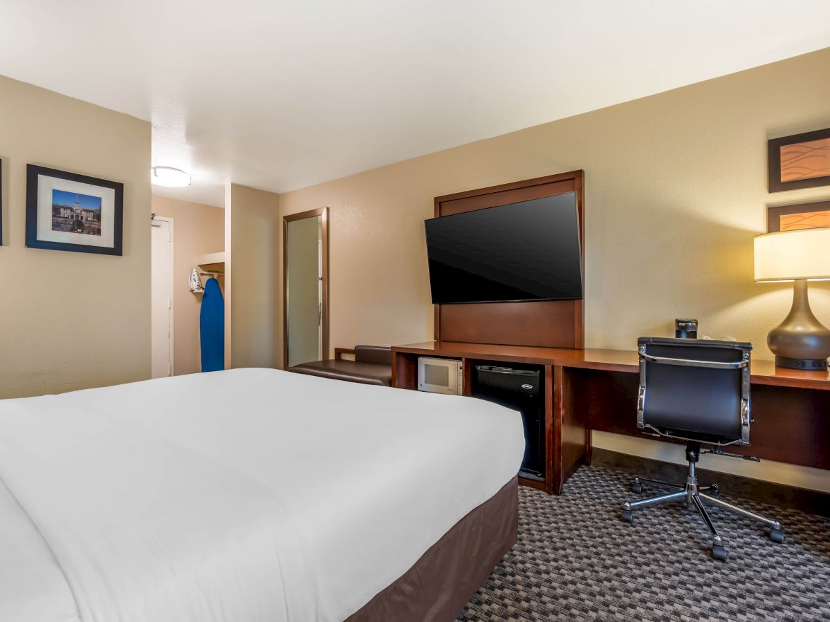 A hotel room with a bed, television, desk, chair, lamp, and blue ironing board in the closet. The walls have two framed artworks.