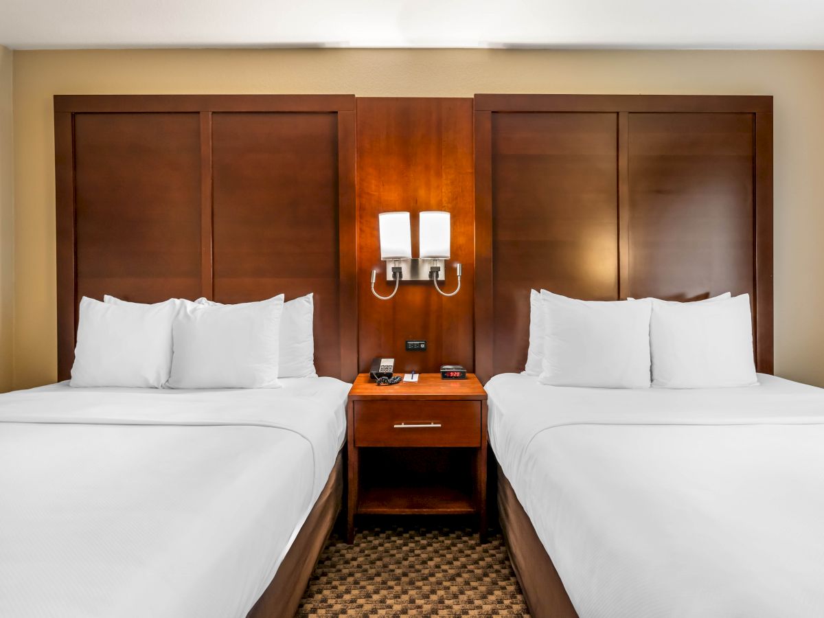 The image shows a hotel room with two neatly made double beds, separated by a wooden nightstand with a lamp, phone, and alarm clock.