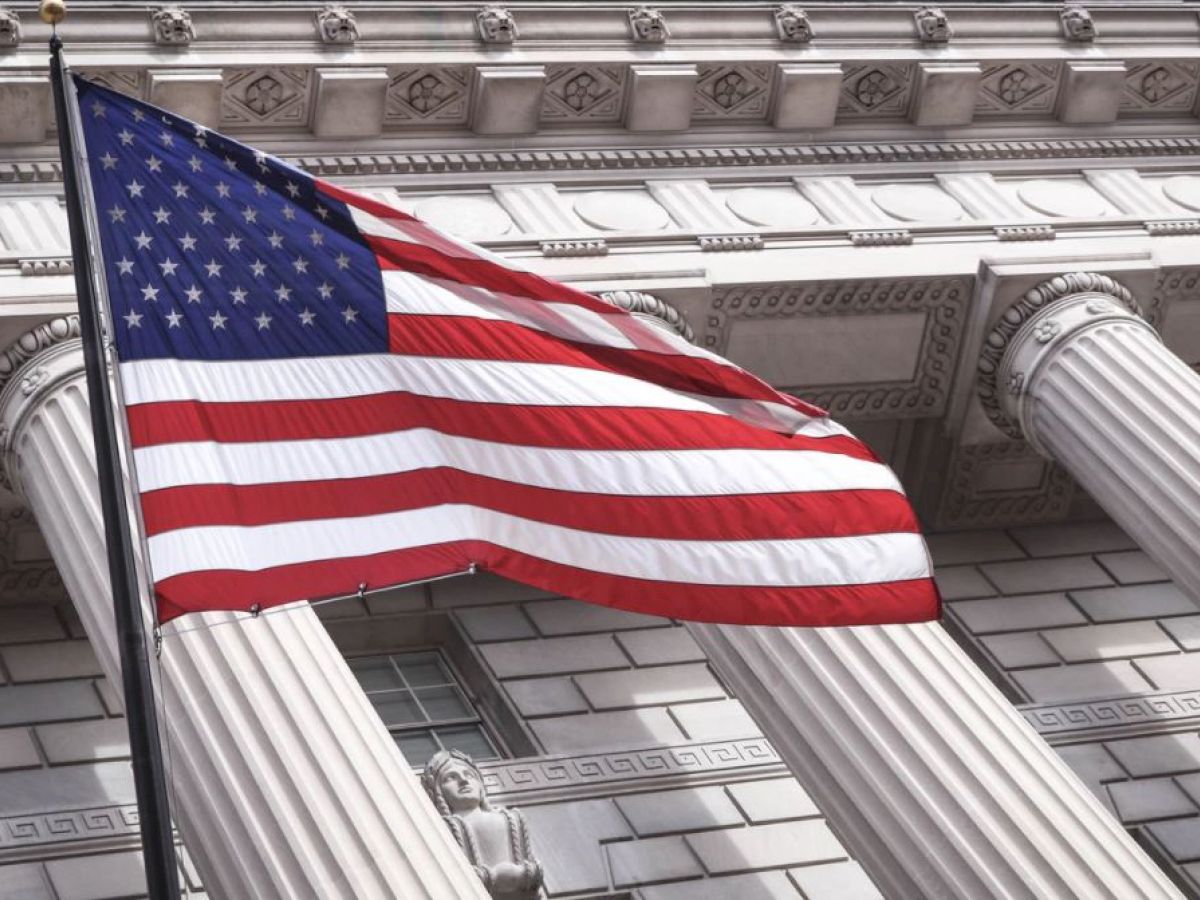 An American flag waves in front of a neoclassical building with large columns and ornate architectural details.