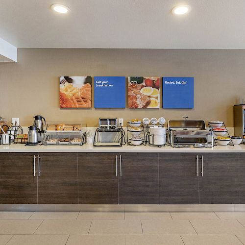 A breakfast buffet setup features cereals, bread, coffee, juice, cooking appliances, and condiments beneath posters displaying food images.