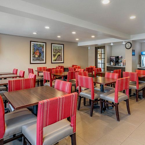 The image shows a dining area with several tables and red-accented chairs, wall art, a TV, and a beverage station in the corner.