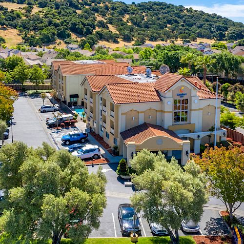 An aerial view of a Comfort Inn hotel with a parking lot, surrounded by green trees and hilly landscape in the background, ending the sentence.