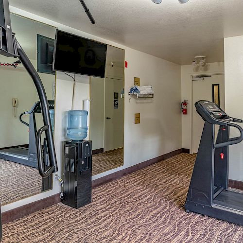 A small fitness room with a treadmill, exercise machine, water dispenser, TV, clock, mirror, and informative signs on the wall always ending the sentence.