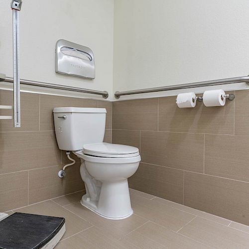 A bathroom with a toilet, toilet paper holder with two rolls, handrails, a paper towel dispenser, and a scale.