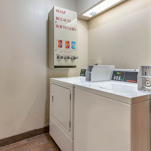 This image shows a small laundry room with a washing machine and dryer, and a vending machine for soap, bleach, and softeners on the wall.