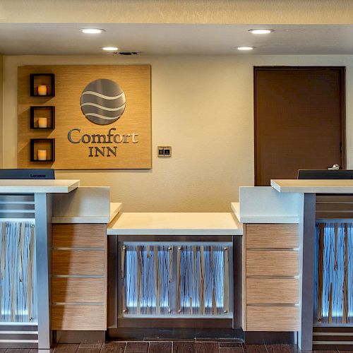 This image shows the reception desk area of Comfort Inn, featuring two desks, a logo on the wall, and a backlit decor beneath the desk.