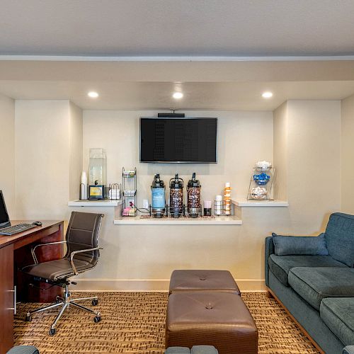 The image shows a cozy lounge area with a computer workstation, comfortable seating, beverage station, TV, and snacks on a shelf under the TV.