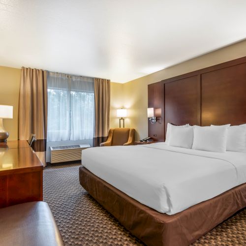 A modern hotel room with a king-sized bed, TV, nightstands, armchair, desk, lamp, and window with curtains, featuring neutral tones and wood accents.