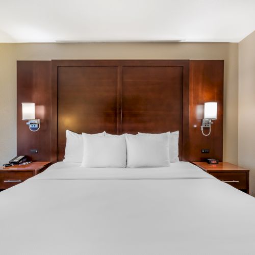 The image shows a neatly made bed in a hotel room with white linens, wooden headboard, two bedside tables, lamps, an armchair, and wall art.