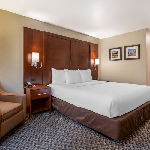 A well-lit hotel room with a large bed, an armchair, bedside tables with lamps, and wall art, featuring neutral tones and a welcoming atmosphere.