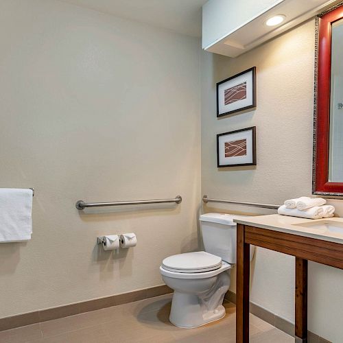 The image shows a clean bathroom with a toilet, sink, towels, a mirror, and wall décor. The shower area is partially visible on the right.