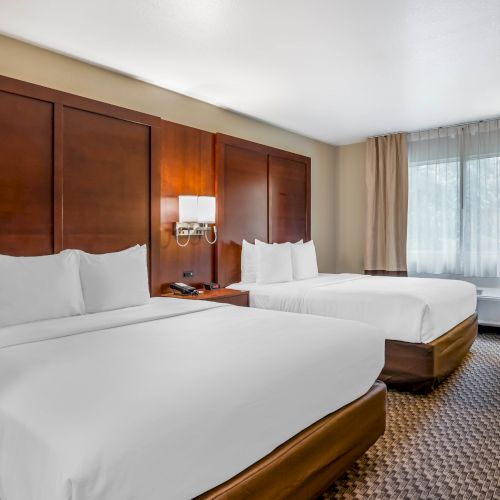 The image shows a hotel room with two beds, white linens, wooden headboards, a nightstand, a window with drapes, and modern decor.