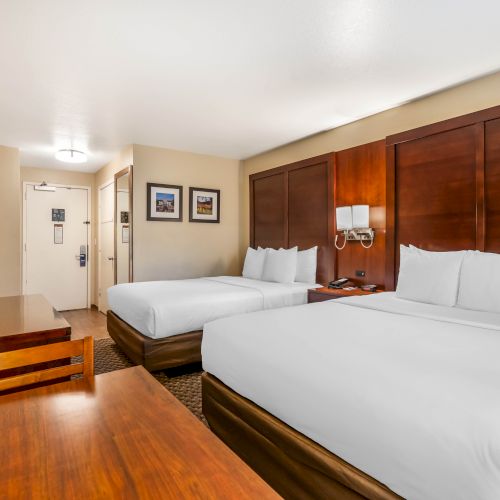 A hotel room with two neatly made double beds, a wooden desk and chair, a wall-mounted TV, and framed pictures on the wall.