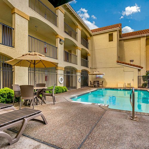 A swimming pool area with lounge chairs, tables, and umbrellas next to a building with balconies under a blue sky with clouds.