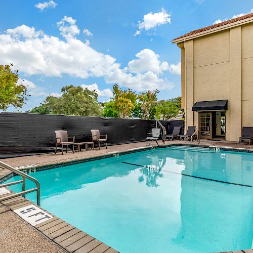 A swimming pool with lounge chairs, umbrellas, and a building in the background on a sunny day.