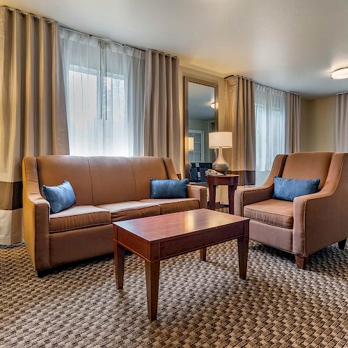 A modern living room features a brown sofa, armchairs, and wooden tables. Gray furnishings and patterned carpet complete the cozy, well-lit space.