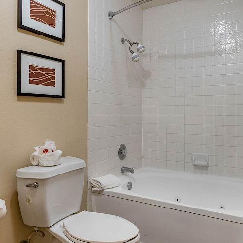 A bathroom featuring a bathtub with a shower curtain, a toilet with folded towels and toiletries, two framed pictures on the wall, and a towel rack.