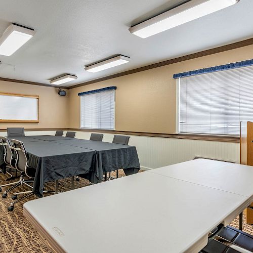 A small conference room with tables, chairs, a podium, and whiteboard, featuring carpeted floors, beige walls, and large windows with blinds.