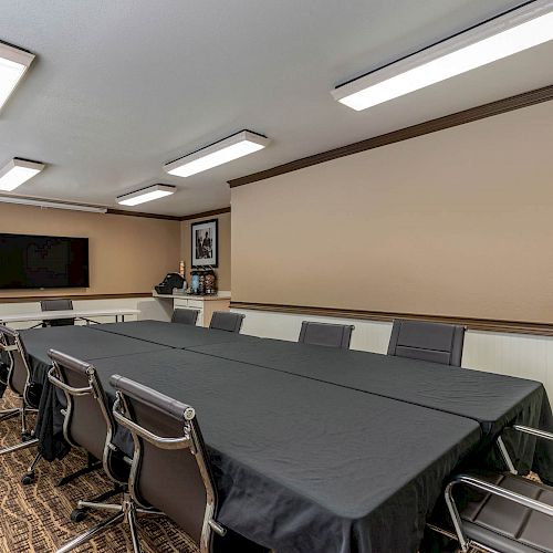 A meeting room with a long black table, multiple chairs, a TV on the wall, and ceiling lights, with a brown carpet and beige walls.