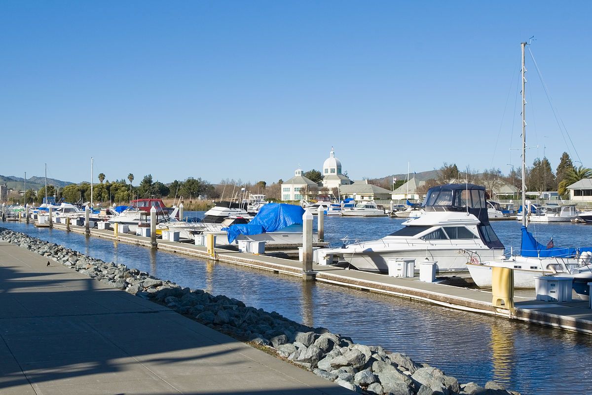 The image shows a marina with numerous boats docked on a sunny day, with a building in the background and a paved walkway along the water.