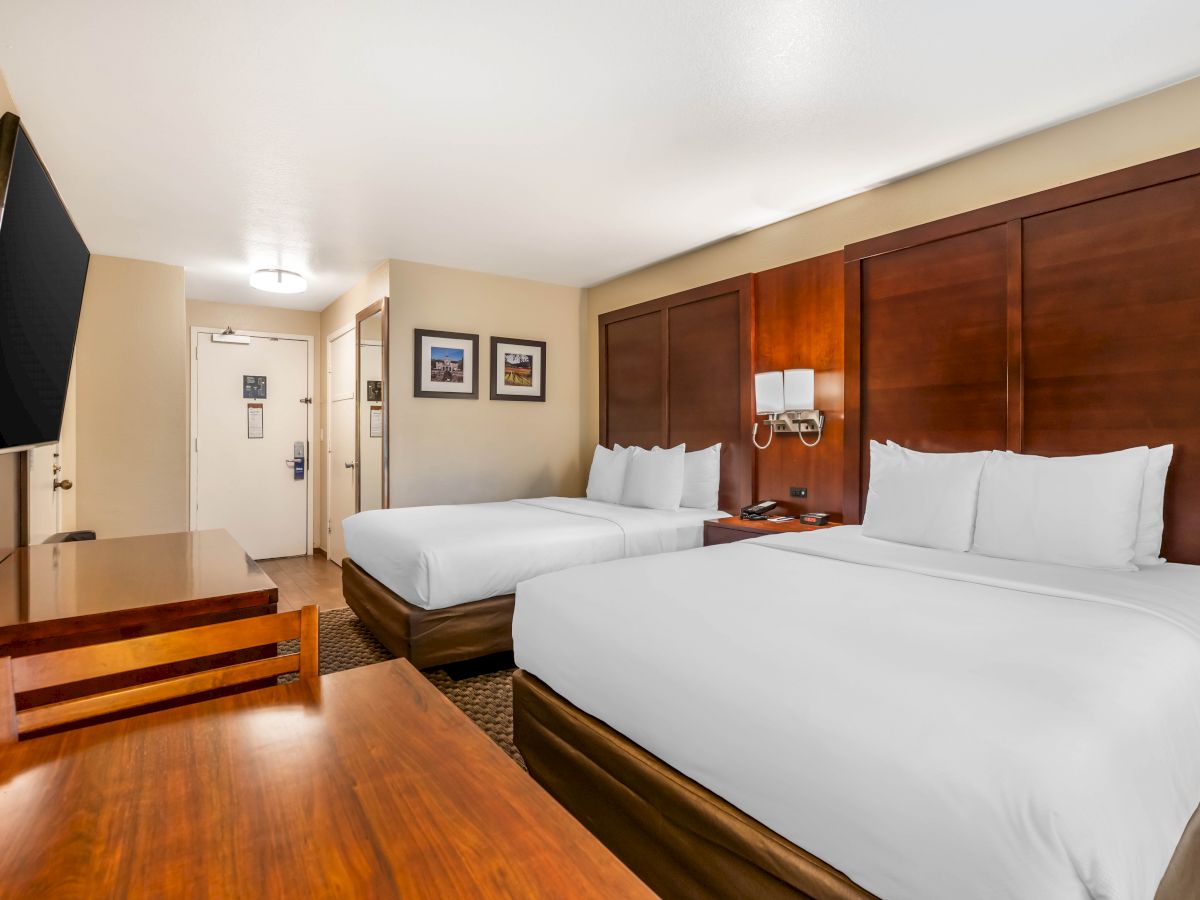 A hotel room with two queen-sized beds, a wood desk, a large TV, and wall-mounted lamps. The entrance and framed pictures are visible.