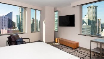 A modern hotel room with large windows offering a city view, a wall-mounted TV, a chair, a small table, and a bed in the foreground.