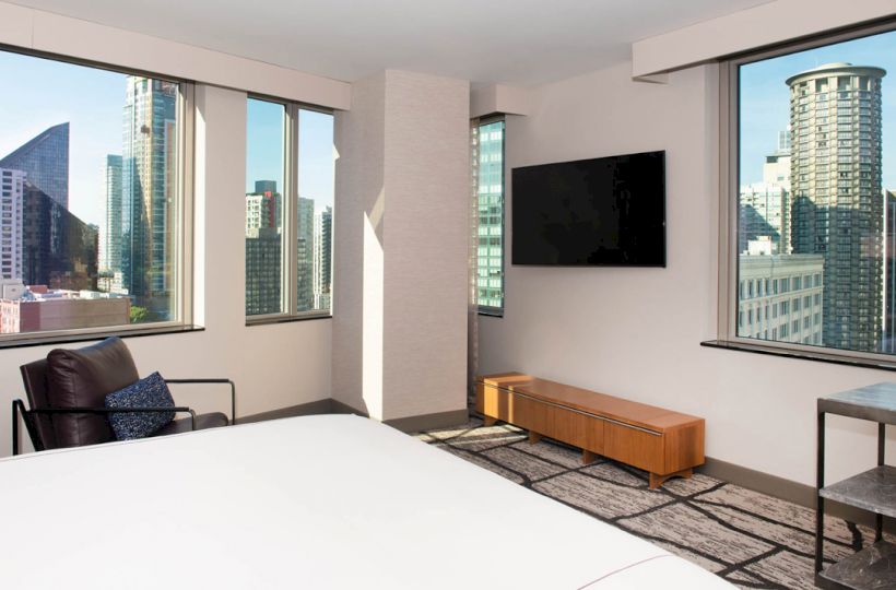 A modern hotel room with large windows offering a city view, a wall-mounted TV, a chair, a small table, and a bed in the foreground.