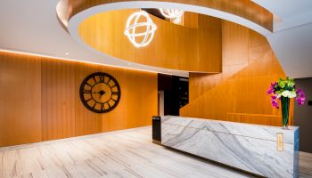 A modern lobby with a marble desk, large clock on the wooden wall, spiral staircase, contemporary lighting and a vase of flowers on the counter.