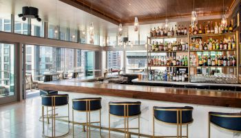 A modern bar with a wooden countertop, blue stools, and numerous bottles displayed on shelves. Large windows offer a cityscape view.