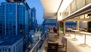 A modern cityscape viewed from a high-rise balcony, with outdoor seating and tables, and large windows revealing an interior space.