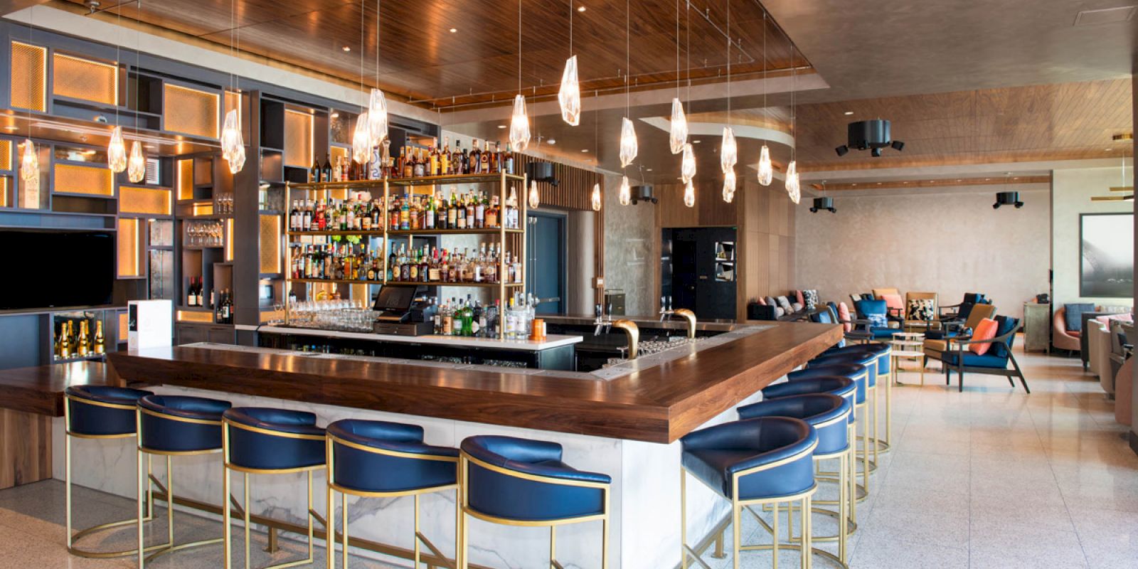 A modern bar with a well-stocked shelf, stylish pendant lights, and blue upholstered bar stools in a beautifully designed interior space.
