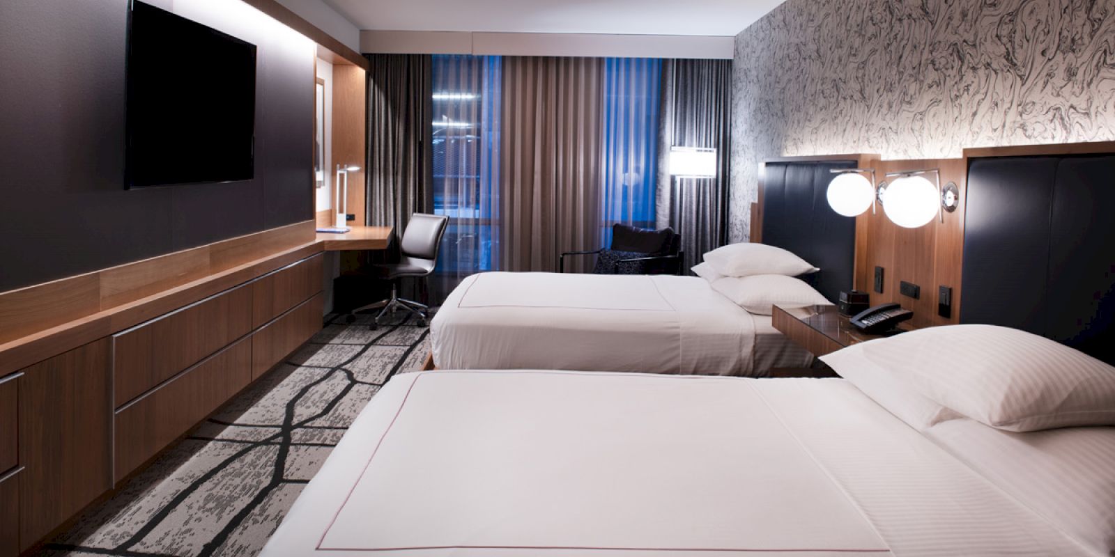 A modern hotel room featuring two neatly made beds, a wall-mounted TV, a desk with a chair, and stylish décor.