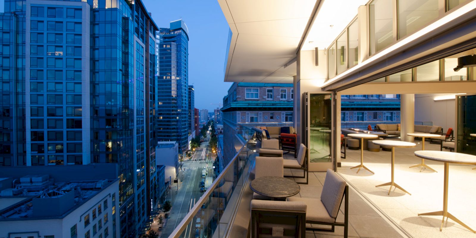 A modern cityscape viewed from a balcony with glass railings, outdoor seating, and a well-lit interior area. The city lights enhance the evening scenery.