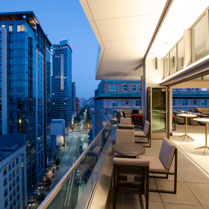 A modern rooftop terrace with seating and tables overlooks a cityscape with tall buildings and a street below at dusk.