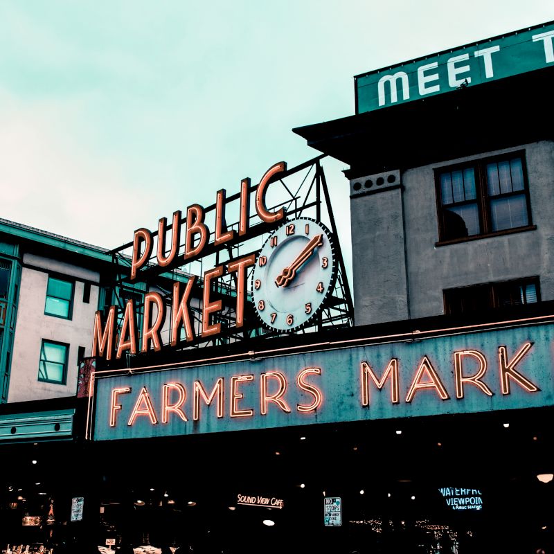 The image shows the exterior of a public market with a prominent neon sign that reads "Public Market" and "Farmers Market" with an old-fashioned clock.