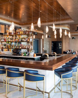 This image shows a modern bar with a wooden counter, blue chairs, hanging lights, and a well-stocked shelf of liquor bottles in the background.