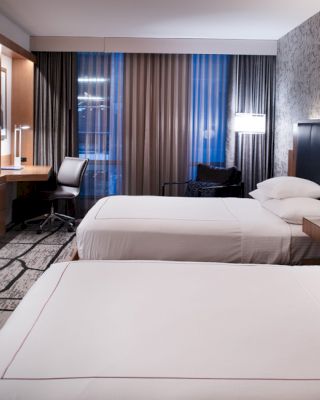A modern hotel room with two double beds, a wall-mounted TV, a desk with a chair, and stylish decor.
