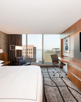 A modern hotel room with a large bed, desk, chair, flat-screen TV, and a view of city buildings through floor-to-ceiling windows.
