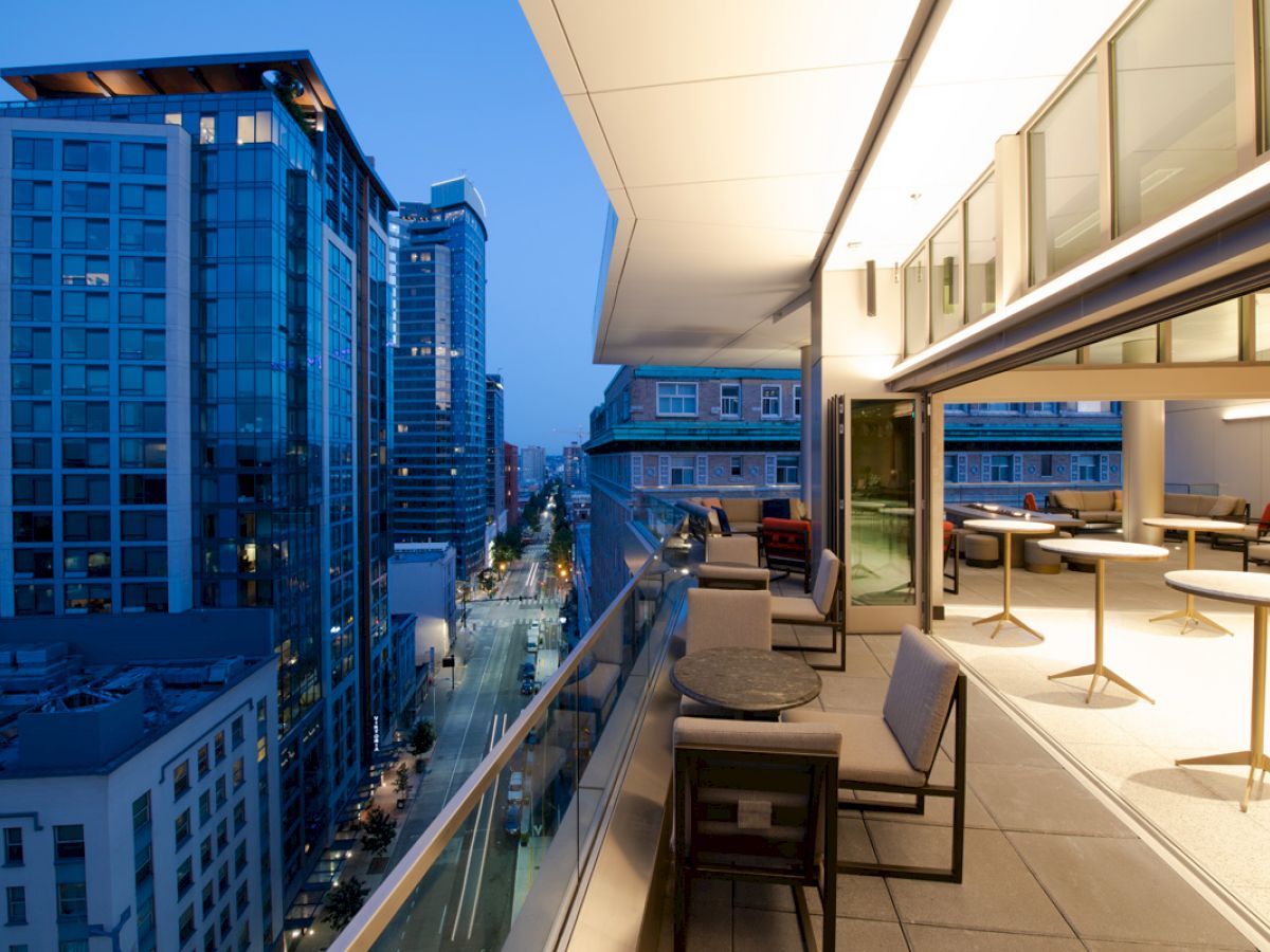 This image shows a modern cityscape viewed from a balcony at dusk, with buildings, streetlights, and outdoor seating on the balcony.