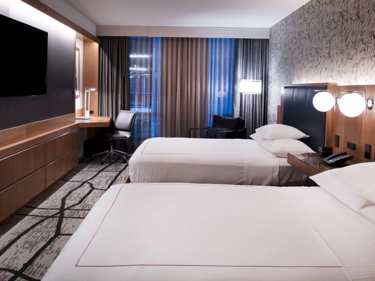 A modern hotel room with two double beds, a wall-mounted TV, desk with chair, and nightstand with lamps. The room has patterned wallpaper.