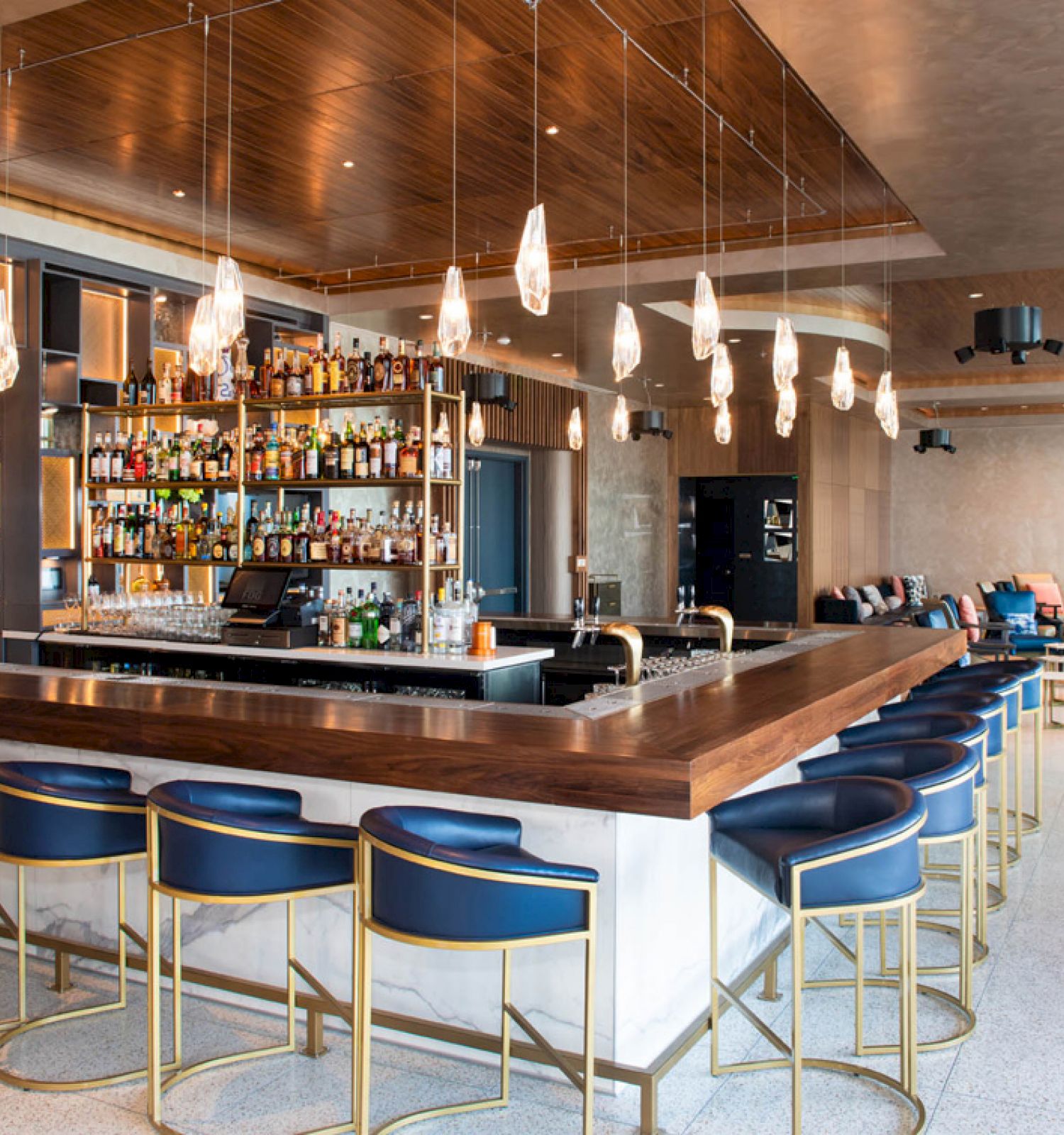 This image shows a modern bar with a central counter, blue barstools, and a well-stocked shelf of bottles behind it. The space is well-lit and stylish.